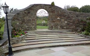 Stonework wall, curved with metal gate installation and steps leading to turfed grass. Designed and built by Evergreen Wales.